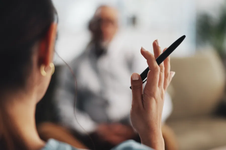 Mujer con un bolígrafo en la mano que explica técnicas de modificación de conductas a un señor al fondo de la imagen sentado en un sofá.